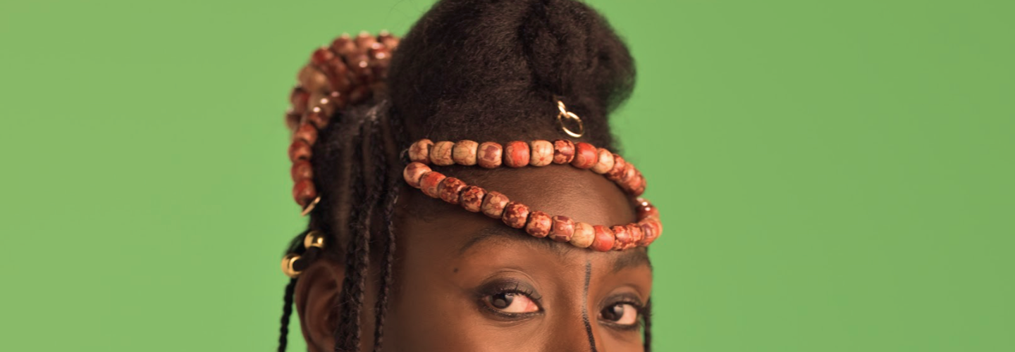 A close-up, cropped shot of a person of color looking directly at the camera. The person has a thick, textured updo with red, clay-like beads woven into the braids and wrapped around the base of the hairstyle. There are small, golden hair accessories interspersed throughout the braids. A thin, black line is drawn from the corner of their left eye down toward their cheek. The background is a solid, bright green.