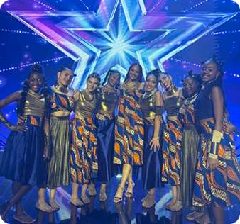 A professional photo of Lil Afro Krew, a group of nine young dancers of various ethnicities, posed on a stage with a large, glowing star in the background. The dancers are dressed in matching outfits featuring pleated gold skirts, and blue and orange patterned tops. They are all barefoot and smiling at the camera.