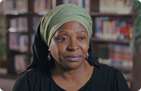 A professional headshot of Dr. Afua Cooper, a Black woman with a warm, gentle smile. She wears a solid, light green headscarf wrapped around her hair, large silver earrings, and a black top. She is seated in front of a bookshelf, suggesting a library or study setting.