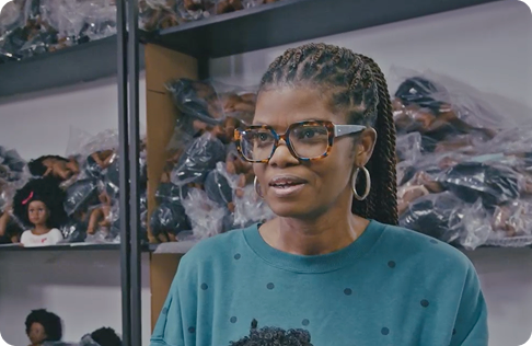 A candid, professional photo of Clara D. Lewis, a Black woman with her hair in neat, braided twists. She is smiling and looking slightly off-camera. She is wearing a dark, teal-colored sweater with black polka dots, large tortoiseshell glasses, and a pair of large silver hoop earrings. She is standing in what appears to be a warehouse or storage room, with shelves of dolls in plastic bags visible behind her.
