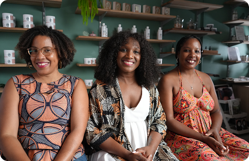 A photo of three smiling Black women, seated side-by-side, from left to right: Catherine Brafine-Dieye, Audrey Belleau, and Annaelle Adaine Jean-Pierre. The woman on the left is wearing a patterned dress with orange, black, and white circular motifs. The woman in the center is wearing a white dress with a patterned, flowing kimono. The woman on the right is wearing a vibrant orange sundress with a tie-dye or floral print. All three are smiling and appear to be in a retail shop with shelves of products in the background.