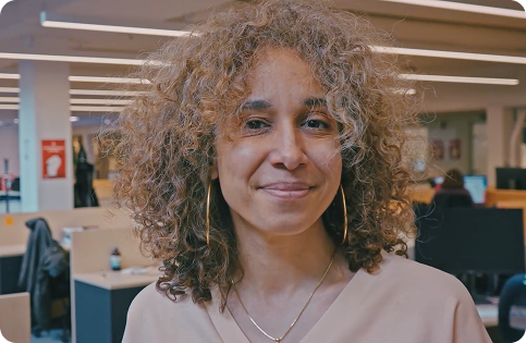 A portrait of a smiling woman with light skin and a mass of curly, light brown hair. She is wearing a light-colored top, thin gold chain, and large gold hoop earrings. She is looking directly at the camera and appears to be in an office environment with desks and computer monitors in the blurred background.