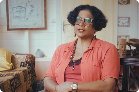 A candid portrait of Juliette Smeralda, a woman with dark, curly hair wearing oversized square-rimmed glasses. She is sitting in a room with a light-colored wall and some decorative artifacts in the background. She is wearing a rust-colored button-up shirt over a lace-trimmed tank top, and a long, multi-strand beaded necklace. She is looking slightly to the side with a serious expression.