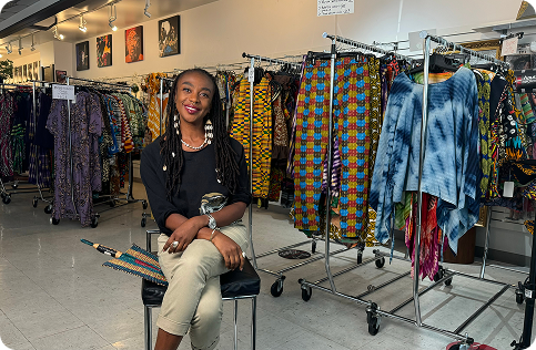 A professional photo of Keithy Antoine, a Black woman with long, braided hair adorned with shells. She is sitting on a chair, smiling at the camera in the middle of a clothing store. She is holding a colorful, woven fan. The store is filled with racks of clothing featuring vibrant patterns and textures.