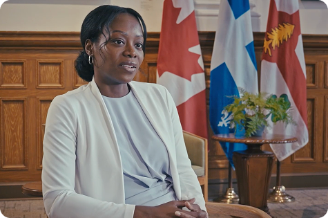 A formal portrait of Martine Musau Muele, a Black woman, seated and looking off-camera. She is wearing a light, cream-colored blazer over a pale grey top and has her hair styled in a low bun. In the background, to the left, is the flag of Canada, and to the right is the flag of Quebec. The setting appears to be a formal office or government building, with polished wood paneling.