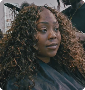 A professional photo of Pascale Lavache, a Black woman, sitting in a chair as she gets her hair braided. She is looking to the side, wearing a protective cape over her shoulders. Her hair is a voluminous mix of loose, brown curls and small, dark braids.