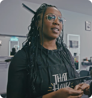 A portrait of a Black woman, Stephanie Odia, wearing glasses and an apron over a black hoodie. She has her dark hair styled in long braids with some strands pulled up in a bun. She is looking off to the side and appears to be in a professional setting, possibly a hair salon, with a chair and mirror visible in the background.